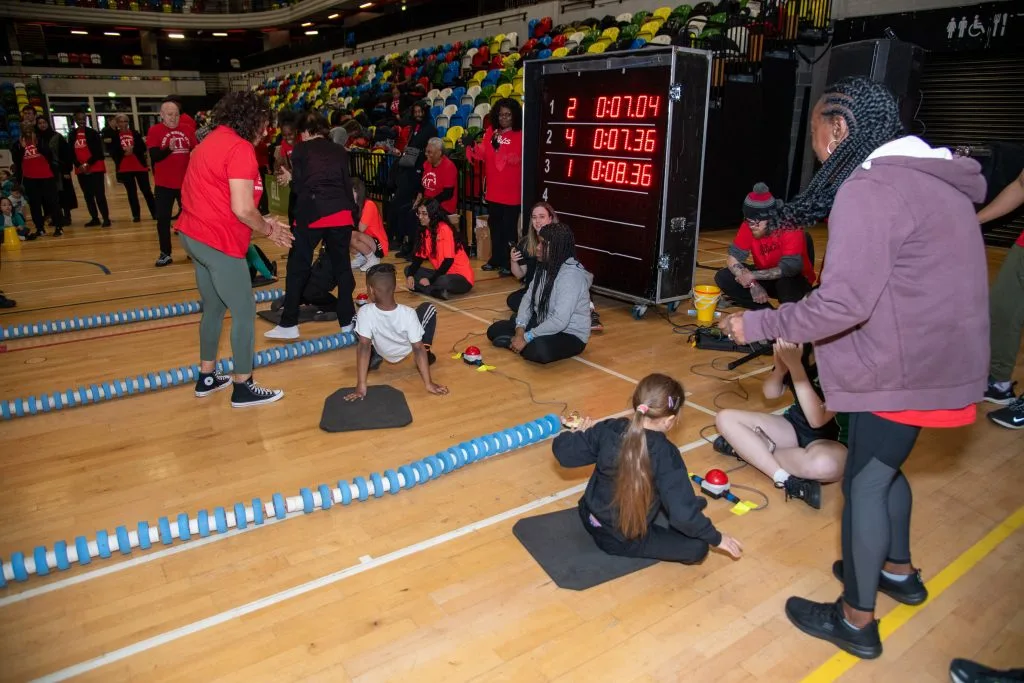 Children on floor mats in a sports hall racing lane, with adults cheering and a digital timing board showing race times