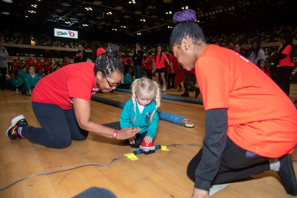 Two adult volunteers kneeling beside a young child on a mat at the start of a race lane