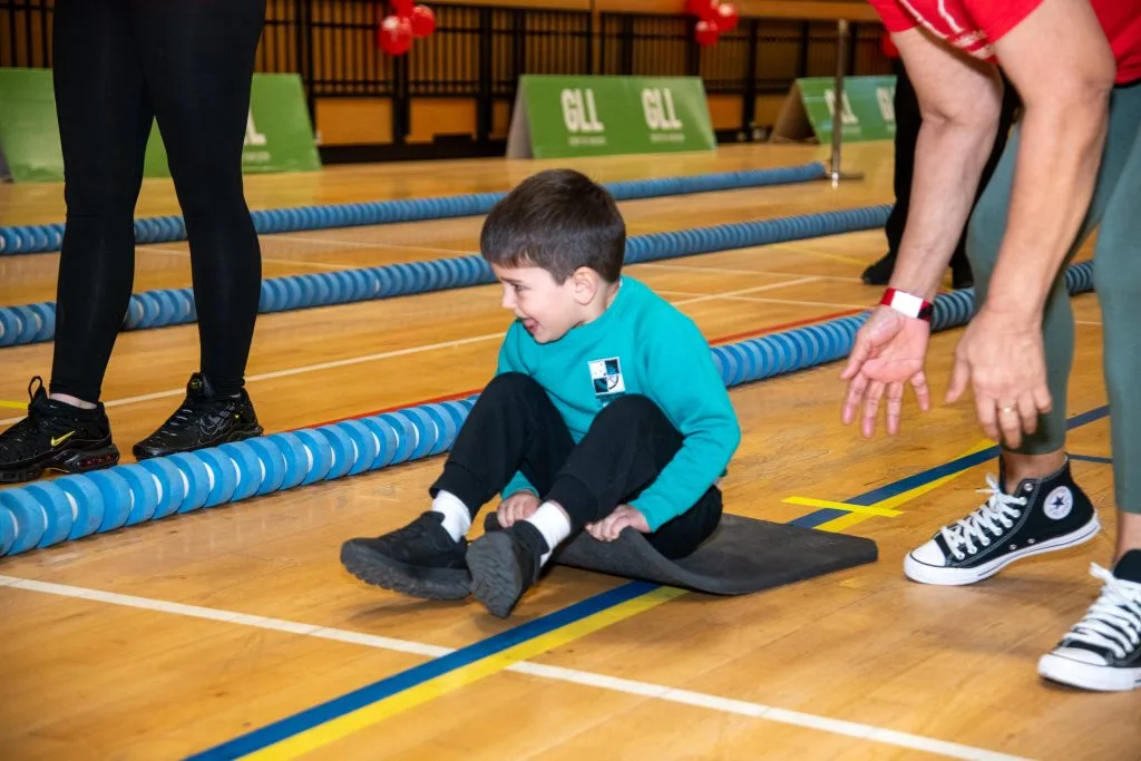 A boy sits on a mat between two floating lane ropes in a sports hall as an adult coach gestures beside him