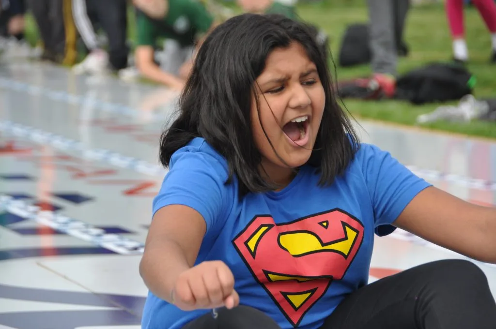 A girl in a Superman T-shirt shouts with effort as she races on a mat outdoors
