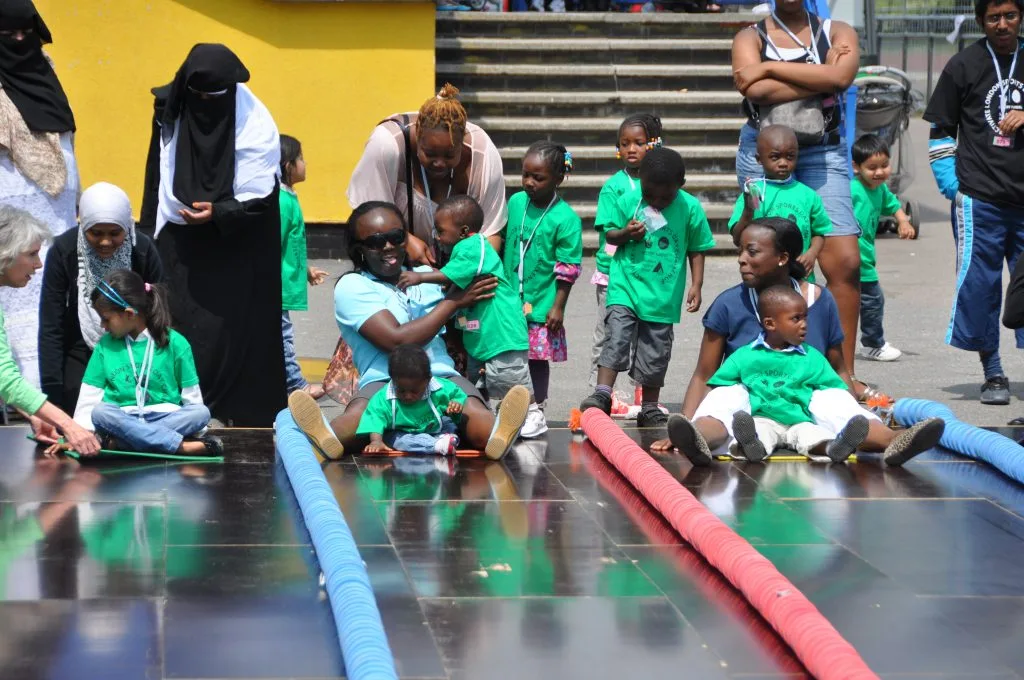 Young children in green T-shirts race on mats between red and blue lane floats in a paved square, watched by parents