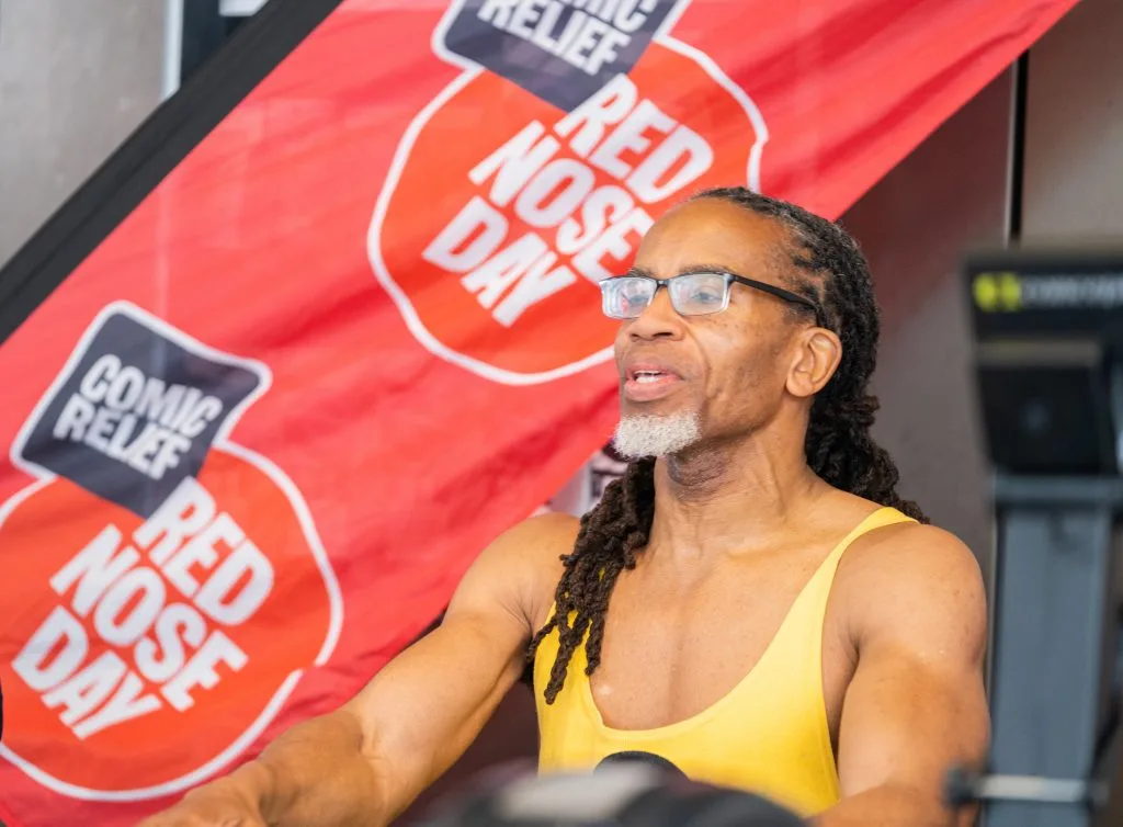 A man in sunglasses and a yellow vest rows in front of a Comic Relief Red Nose Day banner
