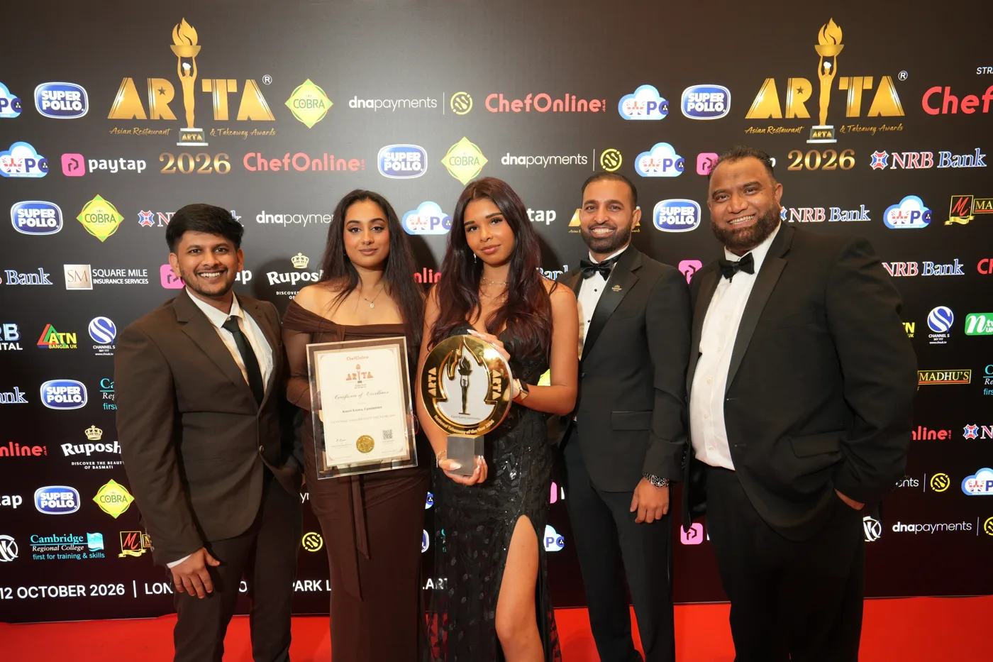 Five members of the Kaani Kaana team in evening dress holding the ARTA trophy and a framed certificate in front of a sponsors' board