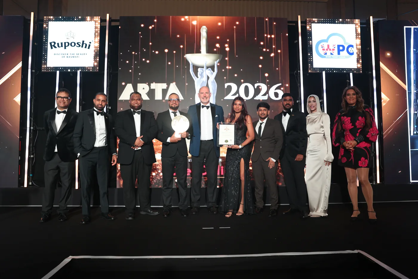 A group of ten people in evening dress on a stage under an ARTA 2026 screen, one holding the trophy and another a framed certificate