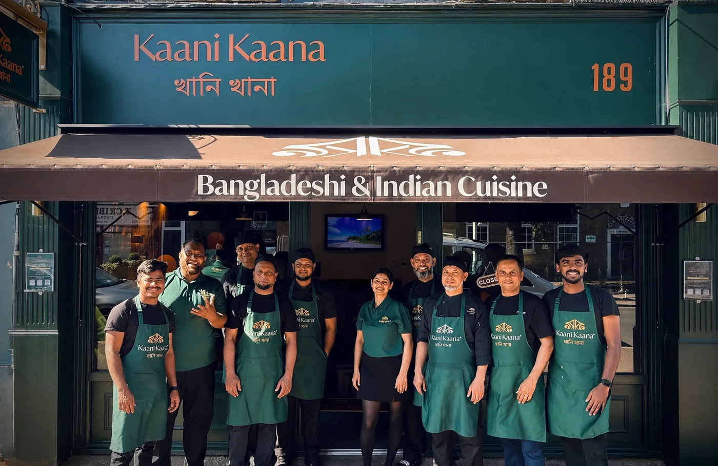 Twelve staff, most in green Kaani Kaana aprons, standing under the restaurant's awning reading Bangladeshi and Indian Cuisine, with the number 189 on the fascia