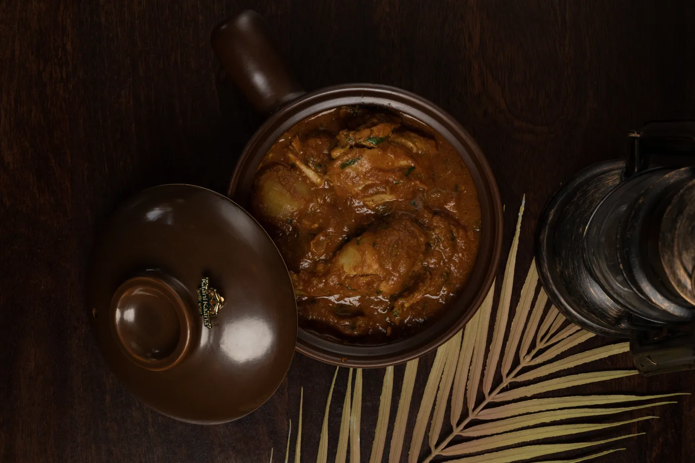 Overhead view of a curry in a brown lidded pot on a dark wooden table beside a lantern and a dried palm leaf