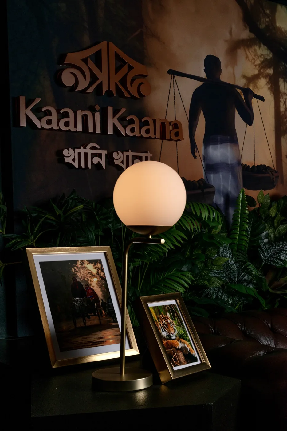 The Kaani Kaana sign in English and Bengali on a dark wall inside the restaurant, above a globe lamp and two framed photographs