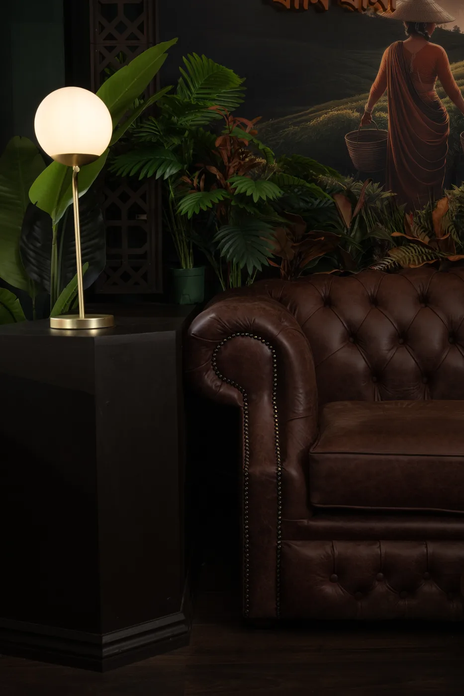 A brown leather Chesterfield sofa beside a globe lamp and plants, under a mural of a woman in a tea field