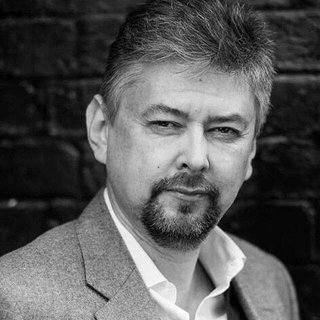 Black and white head and shoulders portrait of Spencer Phillips, a man with short grey hair and a goatee, wearing a jacket and open-necked shirt